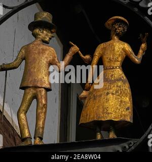 Golden couple.  Gilded centrepiece of an old metal shop sign on an early-18th century timber-framed house in Michelstadt, Hesse, Germany.  The gentleman wears a top hat, has a walking stick hooked over his arm and holds a large tobacco pipe.  The lady wears a summer dress and hat, carries a small handbag in one hand and holds up a bunch of flowers in the other. Stock Photo