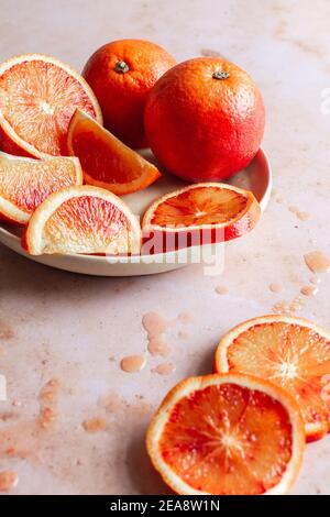Plate of fresh blood oranges, one whole, some halved, sliced or quartered with drops of blood orange juice on the backdrop. Stock Photo