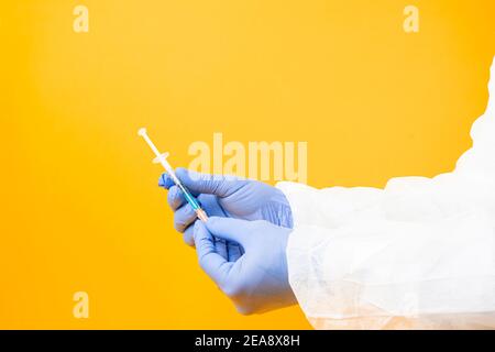 The doctor's hands in protective latex gloves hold a syringe with a loaded needle for intramuscular injection on a yellow background. Vaccination agai Stock Photo