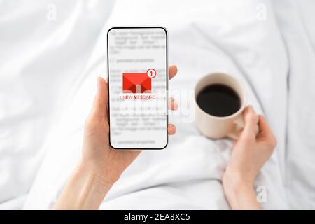 Woman Holding Phone With E-Mail Message Lying In Bed Indoor Stock Photo
