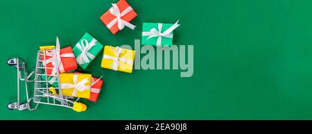 Banner with fallen small cart with scattered gifts. Red, green and yellow gifts in a shopping trolley on a green background. Christmas and Xmas gift c Stock Photo