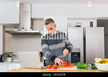 Young man cooking and preparing healthy meal in his kitchen and watching video recipes on the notebook. Preparing healthy food, cutting vegetables Stock Photo
