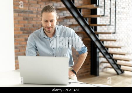 Confident businessman using laptop in loft office space. Friendly male employee sits on the high chair, looks at the screen, researching some tasks Stock Photo