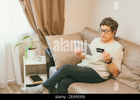 Attractive elegant senior woman sitting on the couch and using a tablet computer and credit card for online shopping at home. Stock Photo