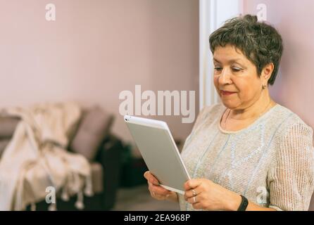 Attractive elegant senior woman staying near the wall and using a tablet computer at home. Stock Photo