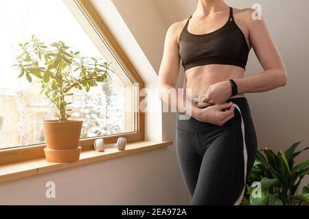 Young beautiful woman measuring waist with tape by a window in attic at sunny day. Healthy living and workout at home. Stock Photo