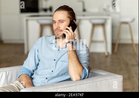 A handsome calm guy talks on the smartphone sitting on the comfortable armchair at stylish apartment. A portrait of businessman during phone Stock Photo