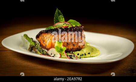 Crispy skin pan seared salmon with peas purée and green garnishes in white plate. Isolated on dark wooden background Stock Photo