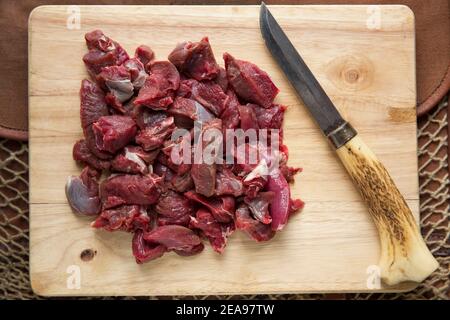 Chunks of raw venison from a wild fallow deer to be cooked in a stew. On a wooden chopping board with a homemade knife. England UK GB Stock Photo
