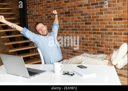 Successful freelancer sits in a confident pose with a feet on the table, raises arms up and stretching, ceo relaxing after good day, a male employee Stock Photo
