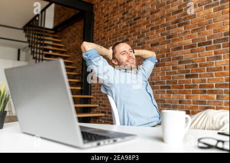 Satisfaction businessman sits in a confident pose with a feet on the table and relaxes, leaned back in the chair, holds his hands behind his head, a Stock Photo