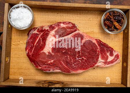 Raw beef ribeye on a round slate plate with glass containers containing peppercorns, coarse salt and cayenne chillies on an old wooden table in brown Stock Photo