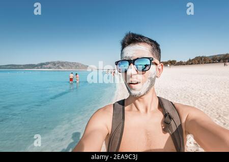 Happy and funny young male tourist in sunglasses takes a selfie at a therapeutic mud resort of Salda lake in Turkey. He applied natural cosmetic clay Stock Photo