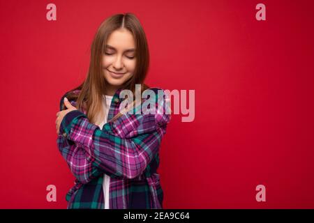 Photo of beautiful cute pretty young blonde woman wearing stylish purple shirt isolated over red background with close eyes and hugging yourself