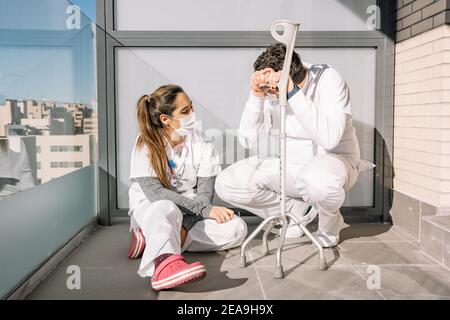 Exhausted male and female medics in masks and uniform sitting on floor on balcony of clinic and taking break during hard working day during coronaviru Stock Photo