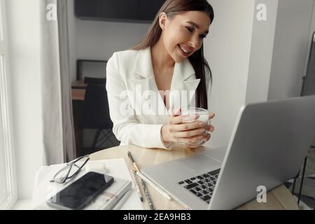 Beautiful happy woman working at home with laptop taking cup of coffee . Smiling business woman woman  in office, using laptop computer Stock Photo