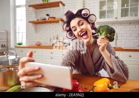 Happy young woman in curlers taking funny selfie with broccoli head in the kitchen Stock Photo