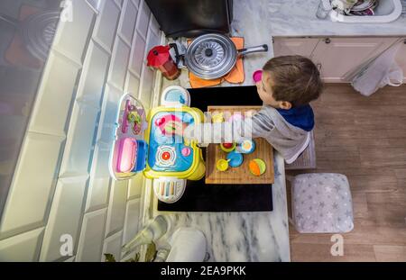 Child boy playing with toy kitchen over real kitchen countertop. Overhead wide angle shot. Slighly motion blurred Stock Photo