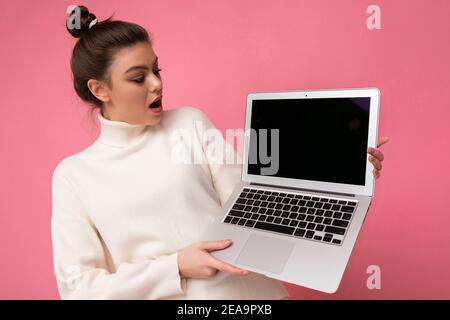 Photo of beautiful amazed and surprised woman with gathered brunette hair wearing white sweater holding computer laptop and looking at the open Stock Photo