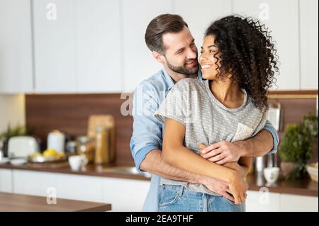 Smiling man hugging from behind charming African American woman, two people standing and joyfully looking at each other. Young international couple Stock Photo