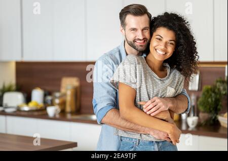 Smiling man hugging from behind charming African American woman, two people standing and joyfully looking at camera. Young international couple Stock Photo