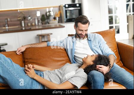 Interracial couple chilling on the couch in the studio apartment, smiling and looking at each other, overjoyed young African American woman lying down Stock Photo