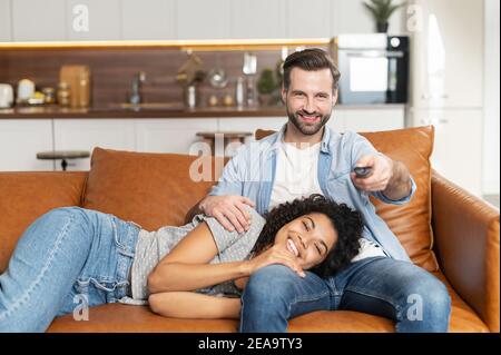Interracial couple chilling on the couch, watching movie in the studio apartment, smiling overjoyed young African American woman lying down on her Stock Photo
