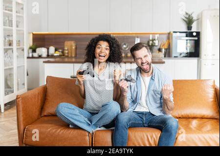 Young African American woman and handsome man sitting on the couch, interracial couple boyfriend and girlfriend playing video game with joysticks in Stock Photo