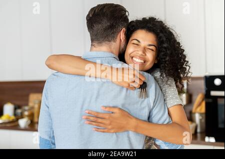 Delighted pleasant young African American woman hugging her boyfriend and feeling thankful for buying a new house, holding keys, and smiling Stock Photo