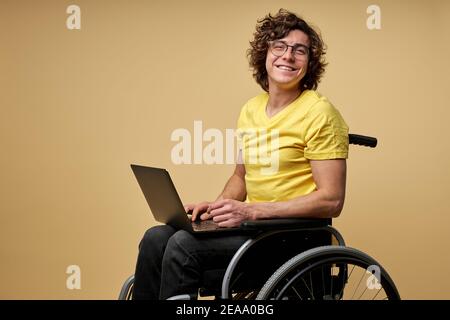 disabled man sits in a wheelchair with laptop, working online. curly young male in casual wear smiles at camera isolated in studio on beige background Stock Photo