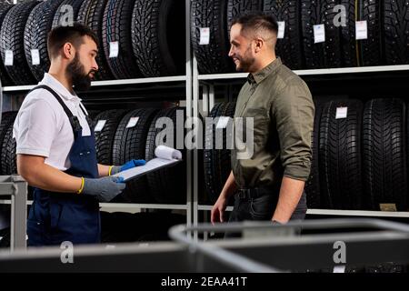 affable auto mechanic in uniform help customer with choice, caucasian young male came to buy new tires for auto. in service Stock Photo