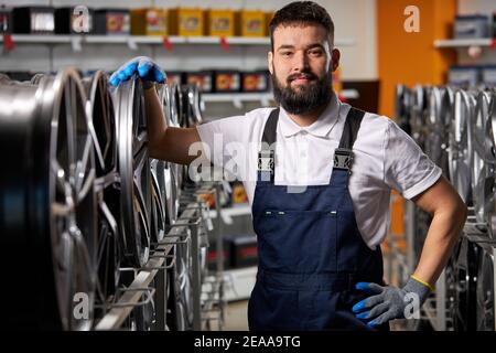 car mechanic in a workshop on a stack of tires at his workplace, young male in uniform stands posing at camera Stock Photo