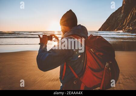 Man travel with backpack taking photo using smartphone camera blogger influencer outdoor walking on beach Stock Photo