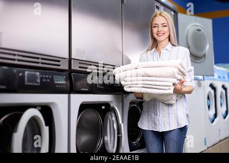 young woman doing laundry at laundromat, looks at camera smiling, holding clean towels in hands and standing near washing machines. washing, cleaning, Stock Photo