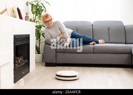 senior woman lying on couch and Robot vacuum cleaner in the room Stock Photo