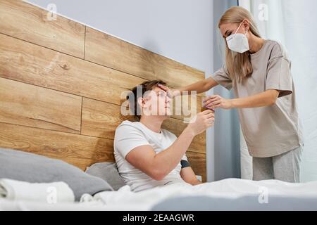 worried wife take care his sick husband while she is sitting on a bed at home checking temperature,people must be wearing medical mask protecting from Stock Photo
