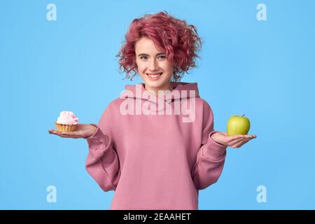 Pretty smiling girl with pink hair in pink hoodie holding fresh green apple and sweet cupcake with cream in hands over blue background. Healthy lifest Stock Photo