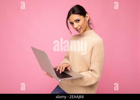 Photo of Beautiful young brunette woman wearing beige sweater holding netbook computer typing text on keyboard looking at camera isolated over pink Stock Photo