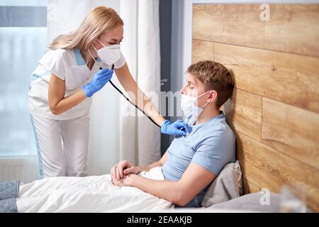 professional female doctor is listening to breath of male patient using stehoscope, at home lying on bed suffering from coronavirus symptoms. covid-19 Stock Photo