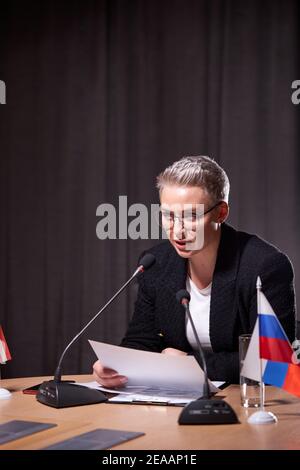 serious female in eyeglasses with short hair sits at business conference using microphone while giving speech to partners. multi-ethnic executives gat Stock Photo