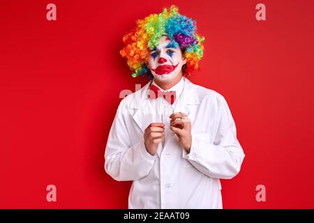 dolorous clown with painted colourful make-up face, upset harlequin isolated over red studio background Stock Photo