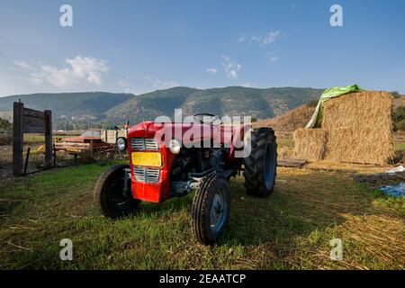 A red old tractor in a countryside farm, surrounded by haystacks, old car, some junk and a wooden sign with mountain ridge in the background. Stock Photo