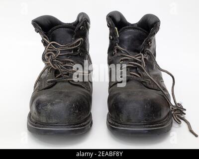 An pair of old, worn, black leather work boots are shown in a front view, isolated against white. Stock Photo