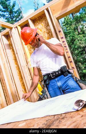 A construction manager talking on a mobile phone while looking at blue print plans on a residential construction site. Stock Photo