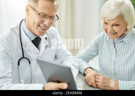 Cheerful doctor using tablet computer while talking with old lady Stock Photo