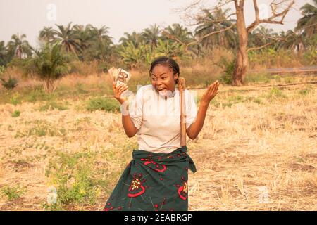 a happy african lady at the farm with a hoe hanging on her shoulder and holding some cash Stock Photo