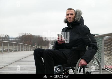 Concept of disabled person. Man in a wheelchair outside in the street. People using technology with smartphone. Stock Photo