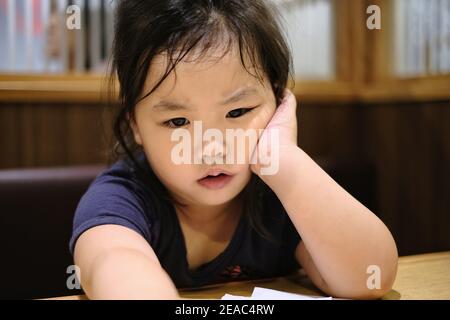 A close up of a cute young Asian girl with an expression of worry and upset on her face, resting her cheek on her hand while resting her elbow on a ta Stock Photo