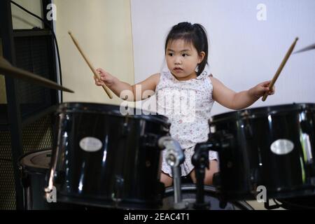 A cute young Asian girl is practicing with her drum set, holding drumsticks in both hands, hitting her drums and cymbals. Stock Photo