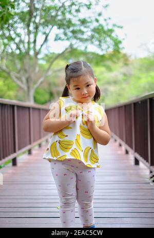 A cute young Asian girl is walking on a wooden bridge over a large pond at a public park in Thailand on a bright sunny day, feeling happy, having fun Stock Photo